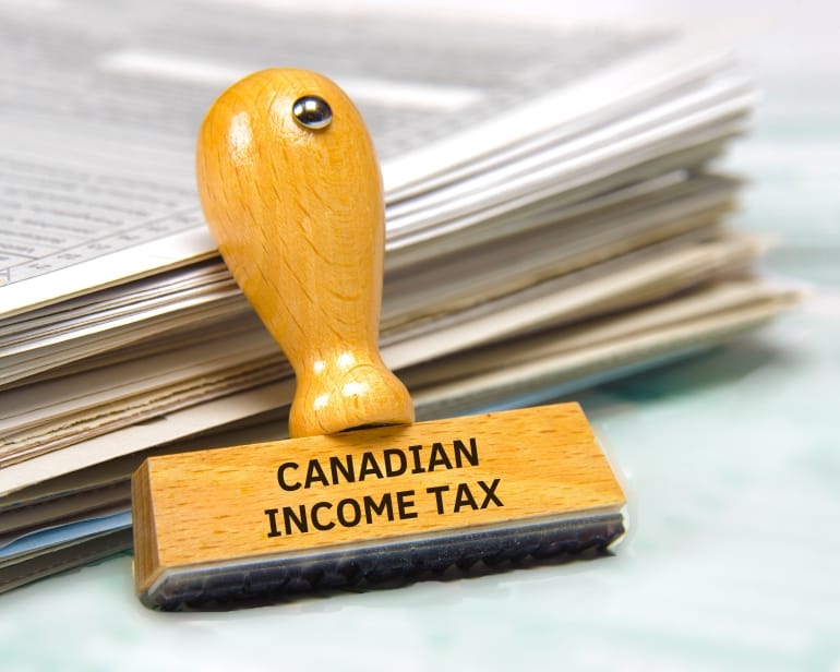 Stack of papers holding up wooden stamp with Canadian Income Tax on it.