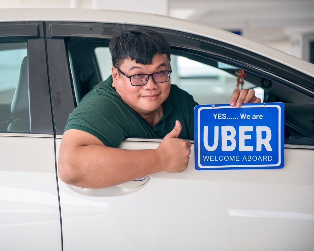 Car with driver leaning out of window with thumb up and holding blue sign which states "Yes..... We are Uber Welcome Aboard.